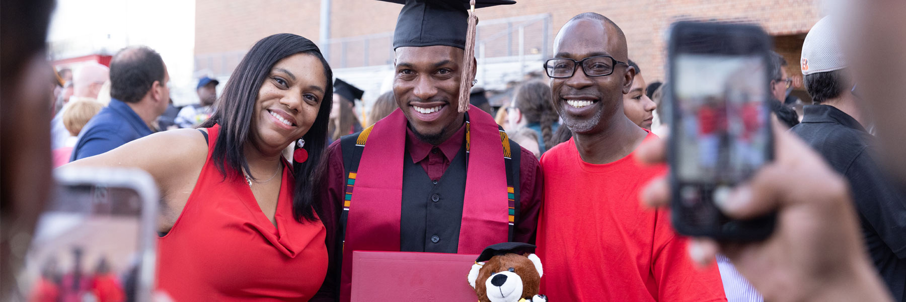 A joyful young man holding his IUE diploma and a teddy bear poses for a family photo with two older relatives 