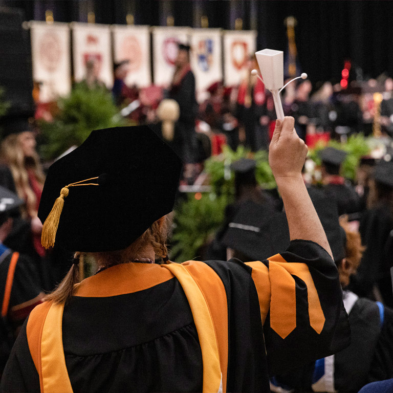 A professor wearing the gold-accented graduation attire of IU's nursing discipline waves a noisemaker during IU East's Commencement ceremony.