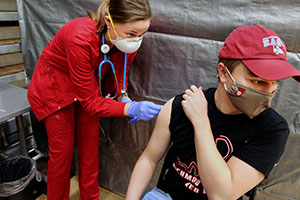 A nursing student in red scrubs looks at a young man while she prepares his arm for a vaccine shot.